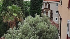 Ein Küstenblick mit zwei hohen Zypressen, einem Gebäude mit rotem Ziegeldach und einer felsigen Insel im Hintergrund.