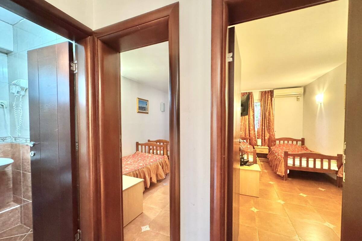 Ein Zimmer mit zwei Betten, einem kleinen Tisch und einem Stuhl. Ein Fenster mit Vorhängen und eine Klimaanlage sind an der Wand angebracht.