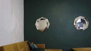 A room with a dark green wall, two hexagonal mirrors, a mustard-colored sofa, and a matching ottoman.