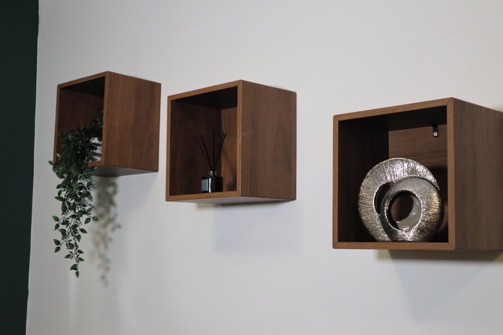 Drei hängende Holzregale mit verschiedenen Gegenständen: eine hängende Pflanze, eine schwarze Vase mit Schilfrohren und ein metallenes, rundes Objekt.