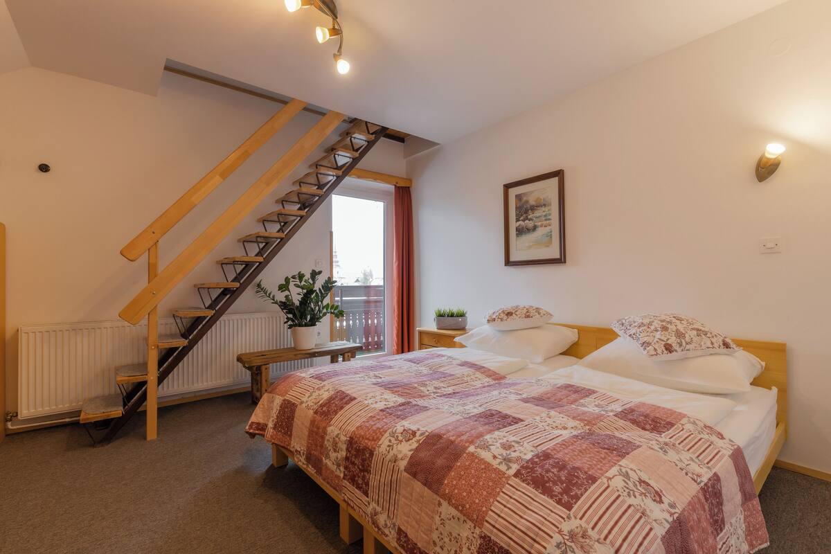 A bedroom with a wooden staircase, a bed with a patterned blanket, a bedside table with a plant, and a window with curtains.