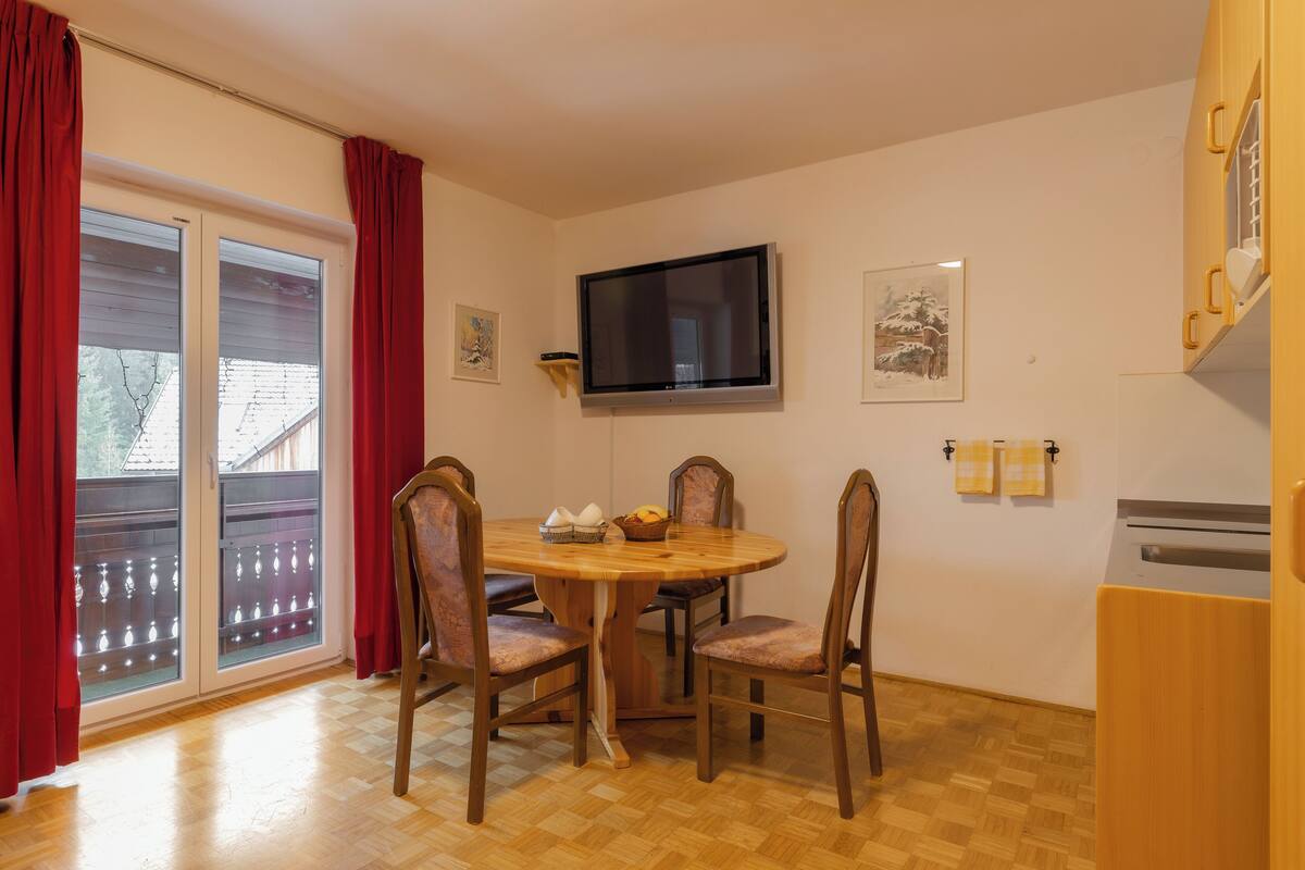 A dining area with a round wooden table, four chairs, a flat-screen TV mounted on the wall, and a window with red curtains.