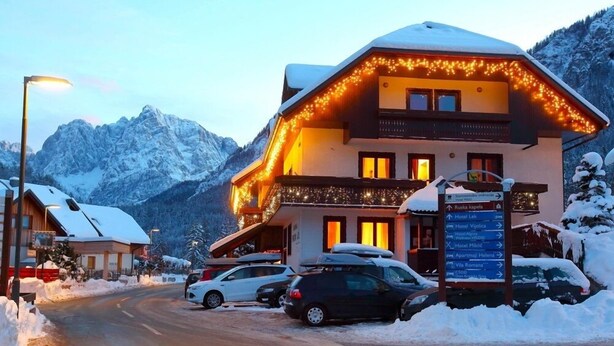 Un hôtel dont le toit est recouvert de neige, un panneau indicateur avec de nombreuses directions, et des voitures garées devant.