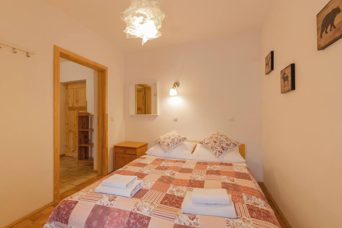 A bedroom with a wooden door, a bedside table, a bed with a quilt, and a wall-mounted light fixture.