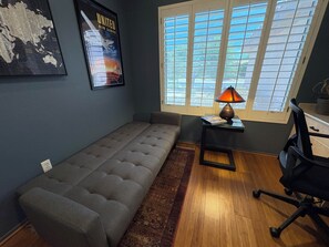 A room with a grey sofa, a small table with a lamp, a window with blinds, and a poster on the wall.