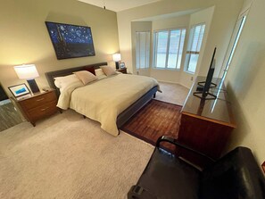 Une chambre d’hôtel avec un lit, des tables de chevet, un bureau avec un ordinateur, une grande fenêtre avec des stores et un tableau au mur.