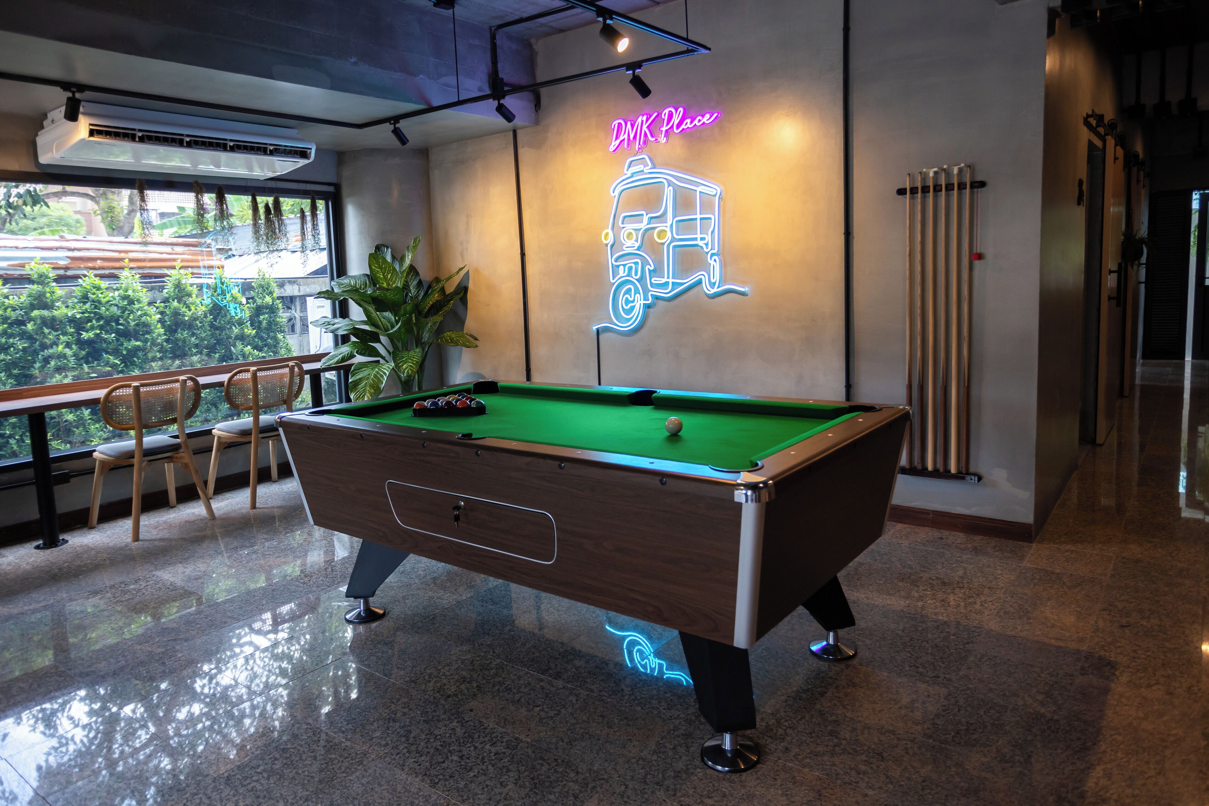 Una mesa de billar en una habitación con un letrero de neón que dice "DMK Place" y vistas a zonas verdes al exterior.