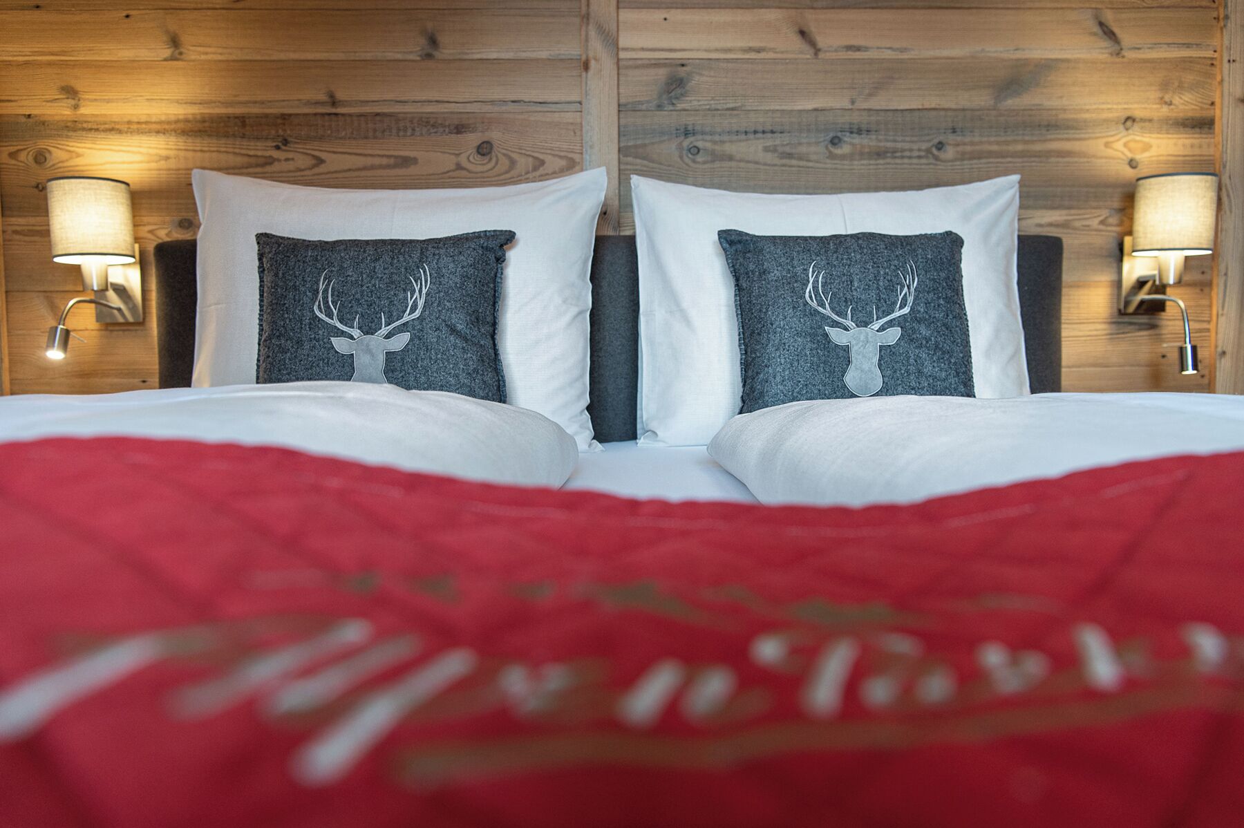 Una cama con una colcha roja y dos almohadas con motivos de ciervo, frente a un cabecero de madera con lámparas fijadas a la pared.