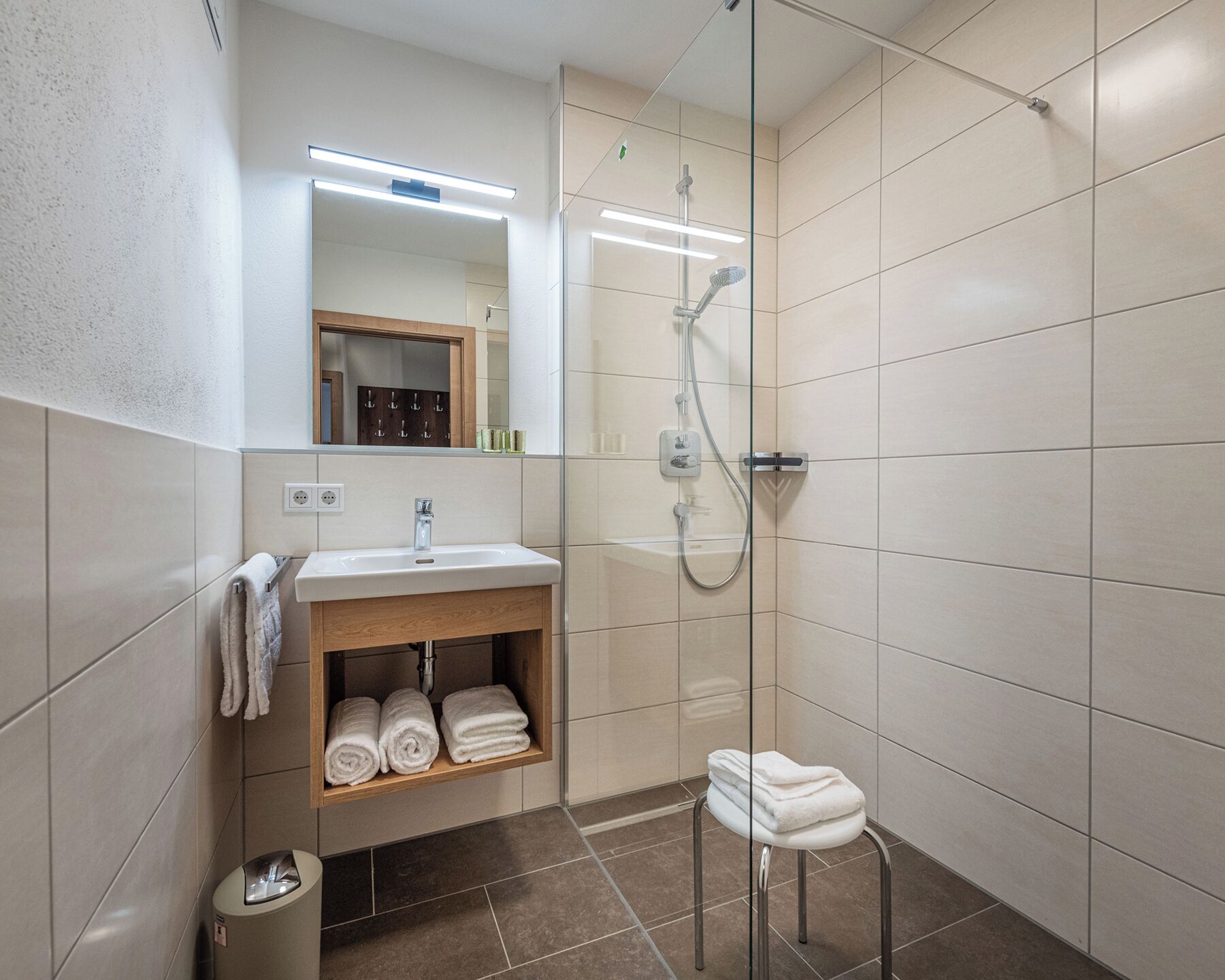 A modern bathroom with a glass-enclosed shower, a sink with a mirror above it, and a shelf with towels.