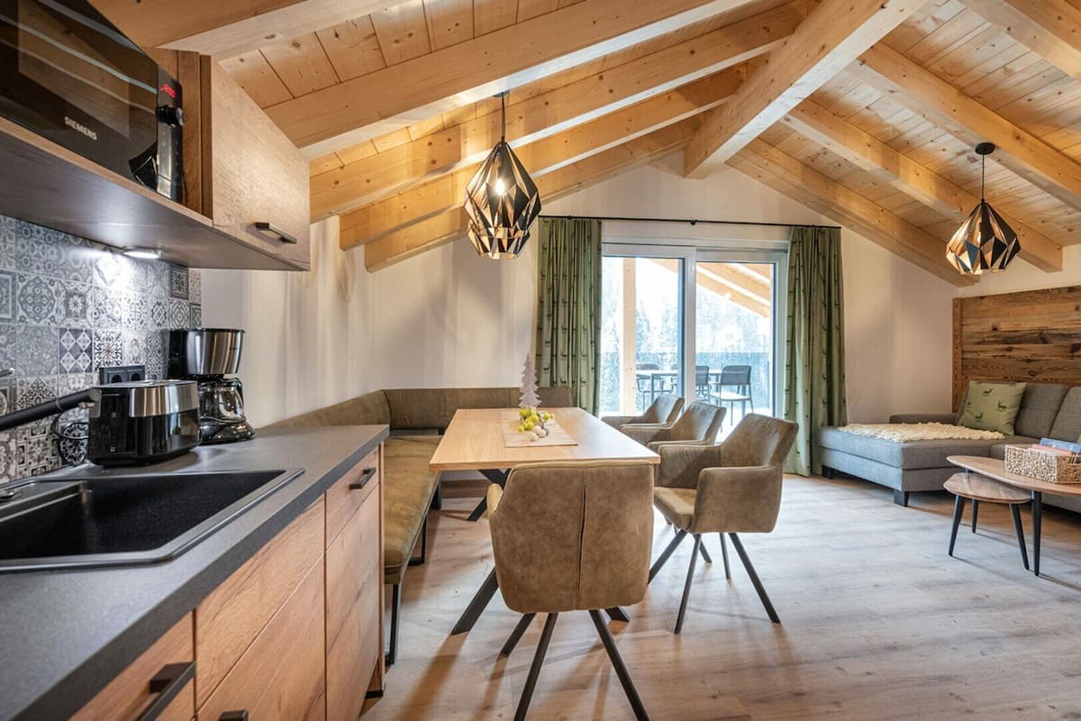 Eine moderne Küche mit Holzbalkendecke, ein Essbereich mit Tisch und Stühlen sowie eine Wohnbereits mit einer Couch und einem Couchtisch.