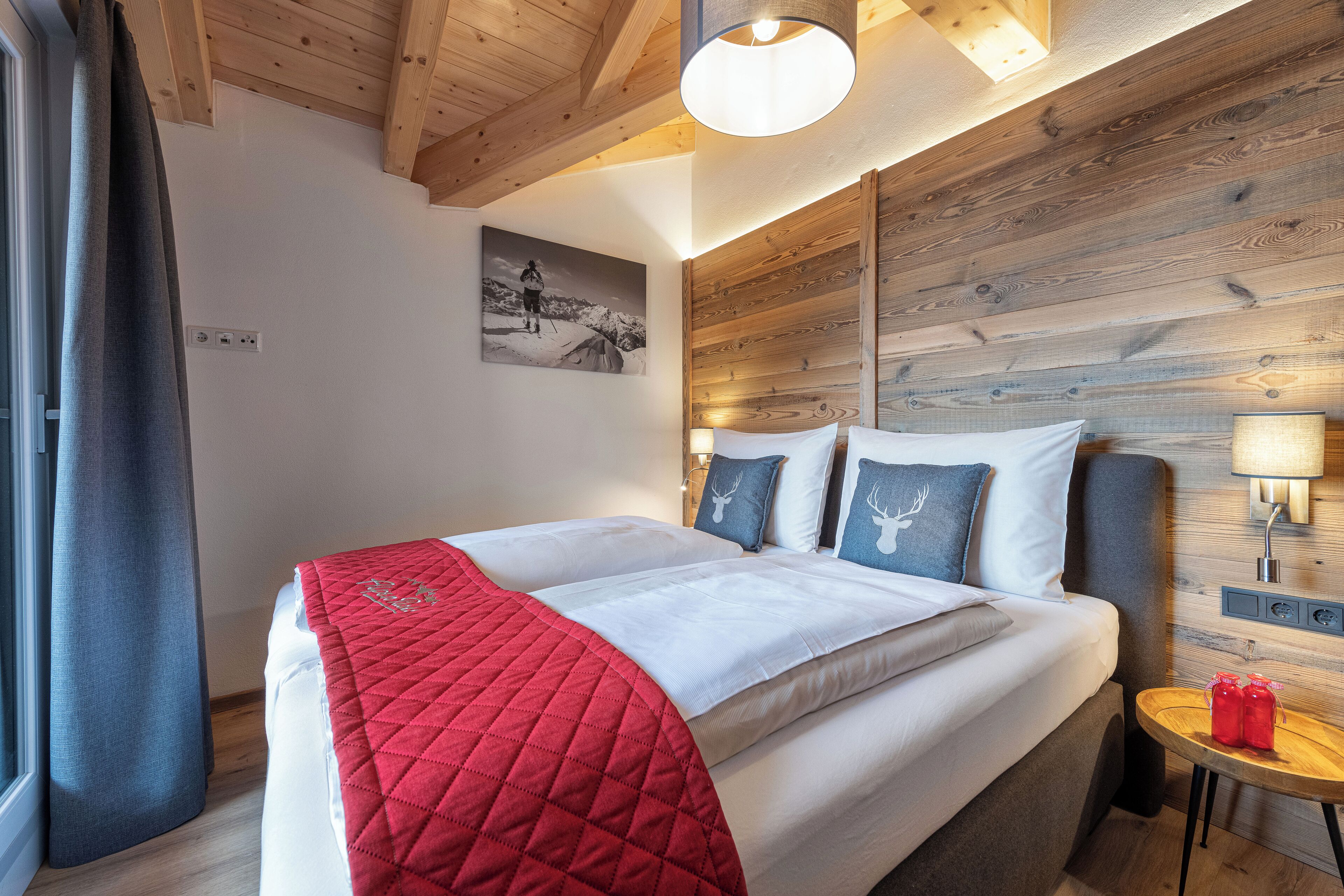 Ein ordentlich bezogenes Bett mit roter Decke, einem hölzernen Kopfteil und einer Wandlampe.