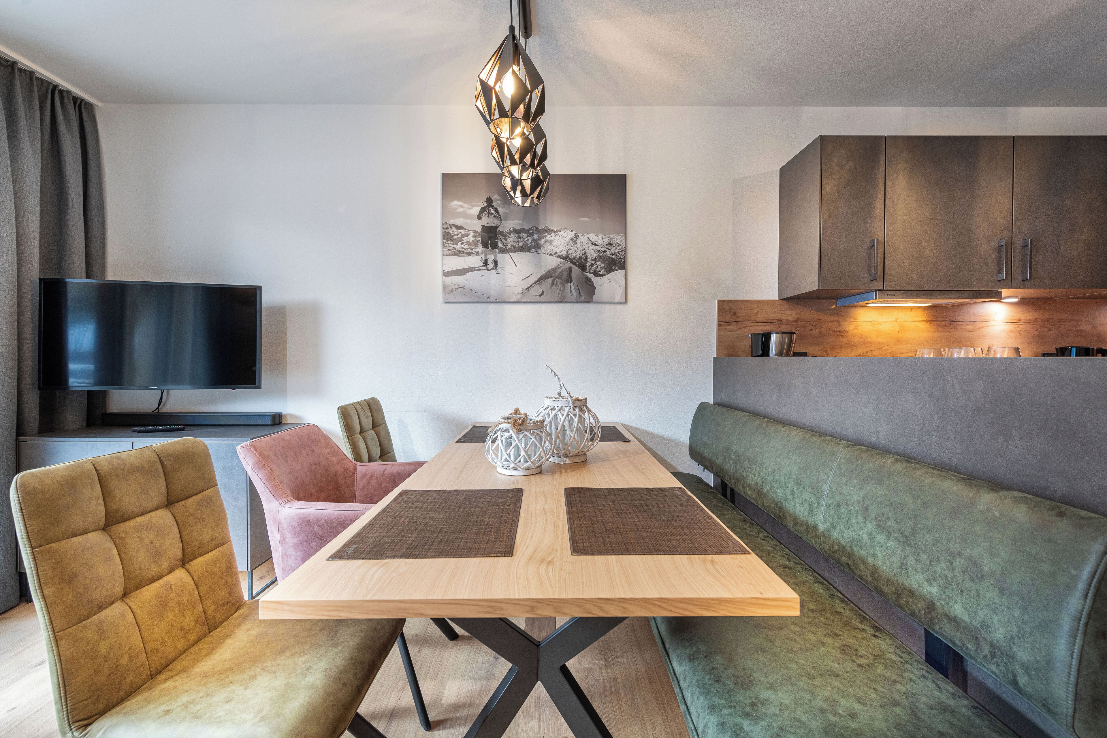 Un salon moderne avec une table à manger en bois, des sièges confortables, un téléviseur à écran plat et un coin cuisine doté de placards et d’un luminaire suspendu.