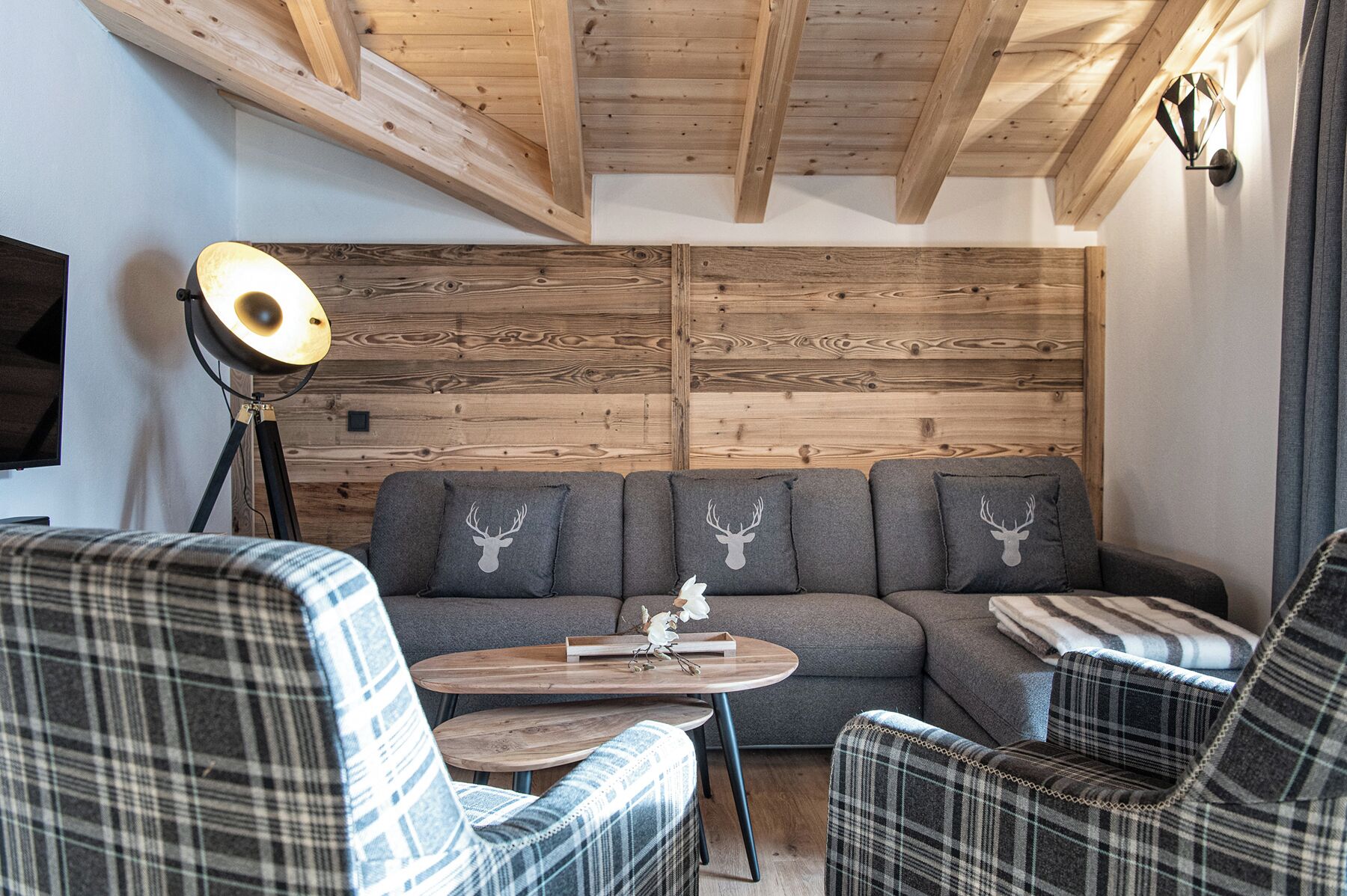 A cozy living room with a wooden ceiling, a sofa with deer head pillows, plaid chairs, and a small wooden table with a decorative item.