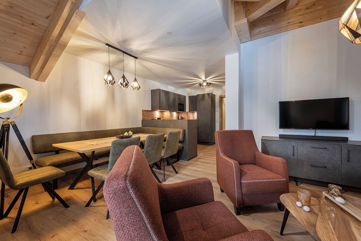 Ein modernes Wohnzimmer mit Holzbalkendecke, einem Essbereich mit langem Tisch und Stühlen, einer Couch, einem Fernseher und einer Küchenzeile mit Schränken und einer Mikrowelle.