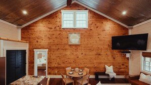 A cozy cabin interior with a wooden interior, a dining table with chairs, a sofa, a television mounted on the wall, and a large window at the top.