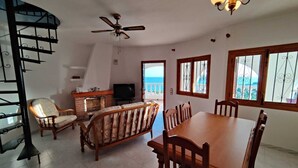 A spacious living area with a fireplace, a TV, and a dining table with chairs. There is a staircase with a railing, a ceiling fan, and a view of the ocean through the windows. - Villa with stunning sea view, private pool and garden (Alcocéber)