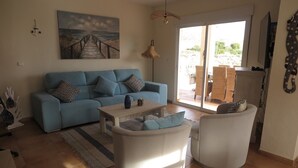 A living room with a blue sofa, a coffee table, and two white armchairs. There is a painting of a beach on the wall, a hanging light fixture, and a view of the outside through a glass door. - beautiful villa with sea and mountain views. (El Campello)