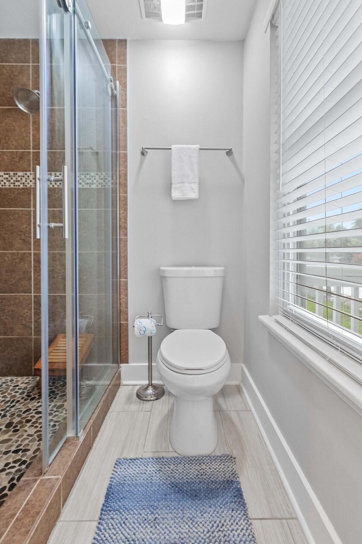 Une salle de bain avec une douche à parois de verre, des toilettes et une fenêtre avec des stores.
