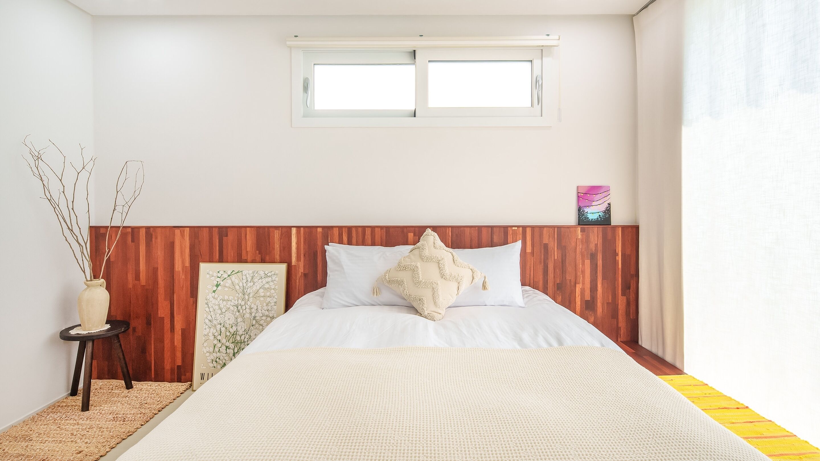 A bedroom with a wooden headboard, a large bed with white bedding, a small side table with a vase, and a window above the bed.