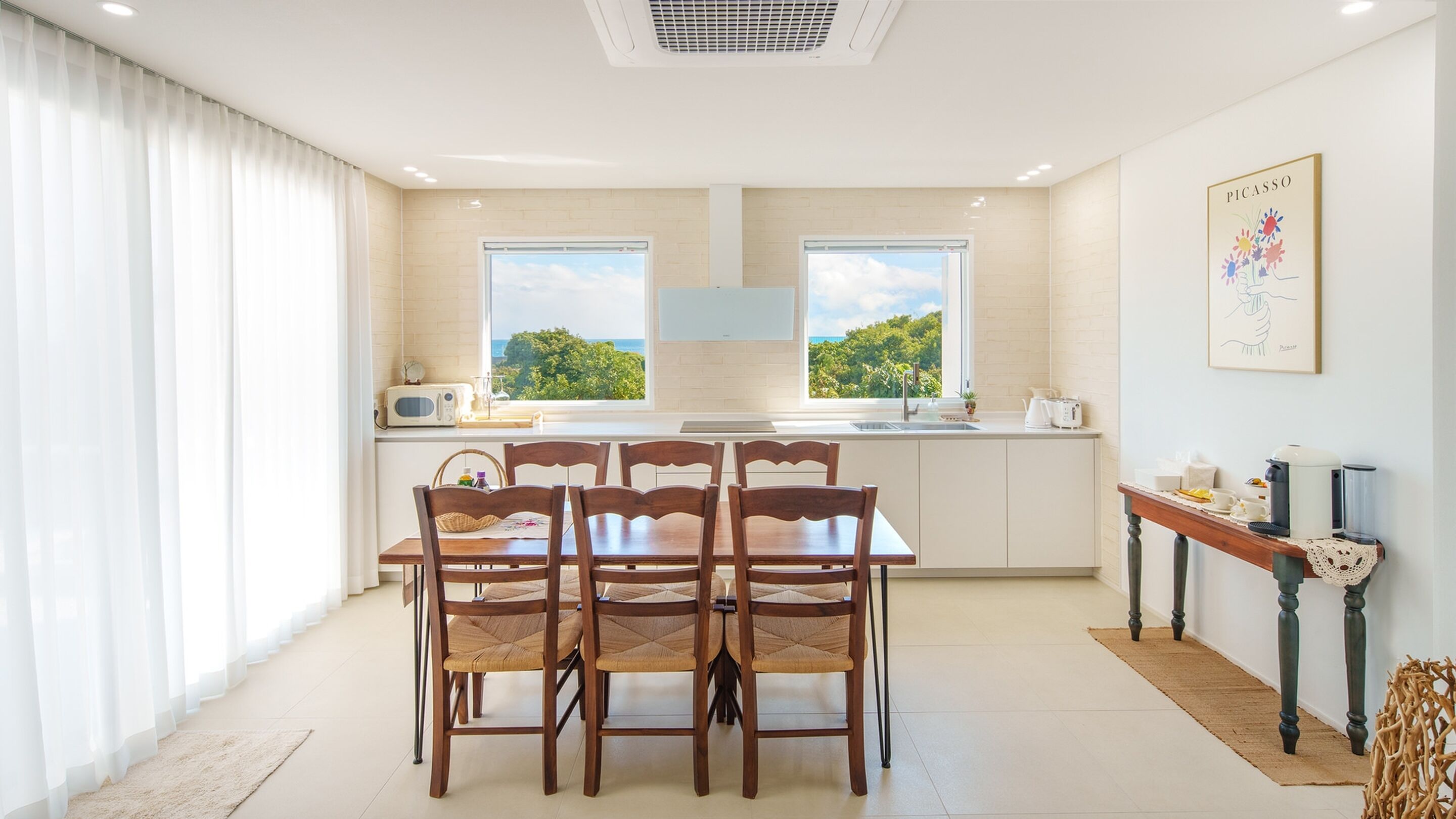 A modern kitchen with a dining area, large windows, and a view of greenery.