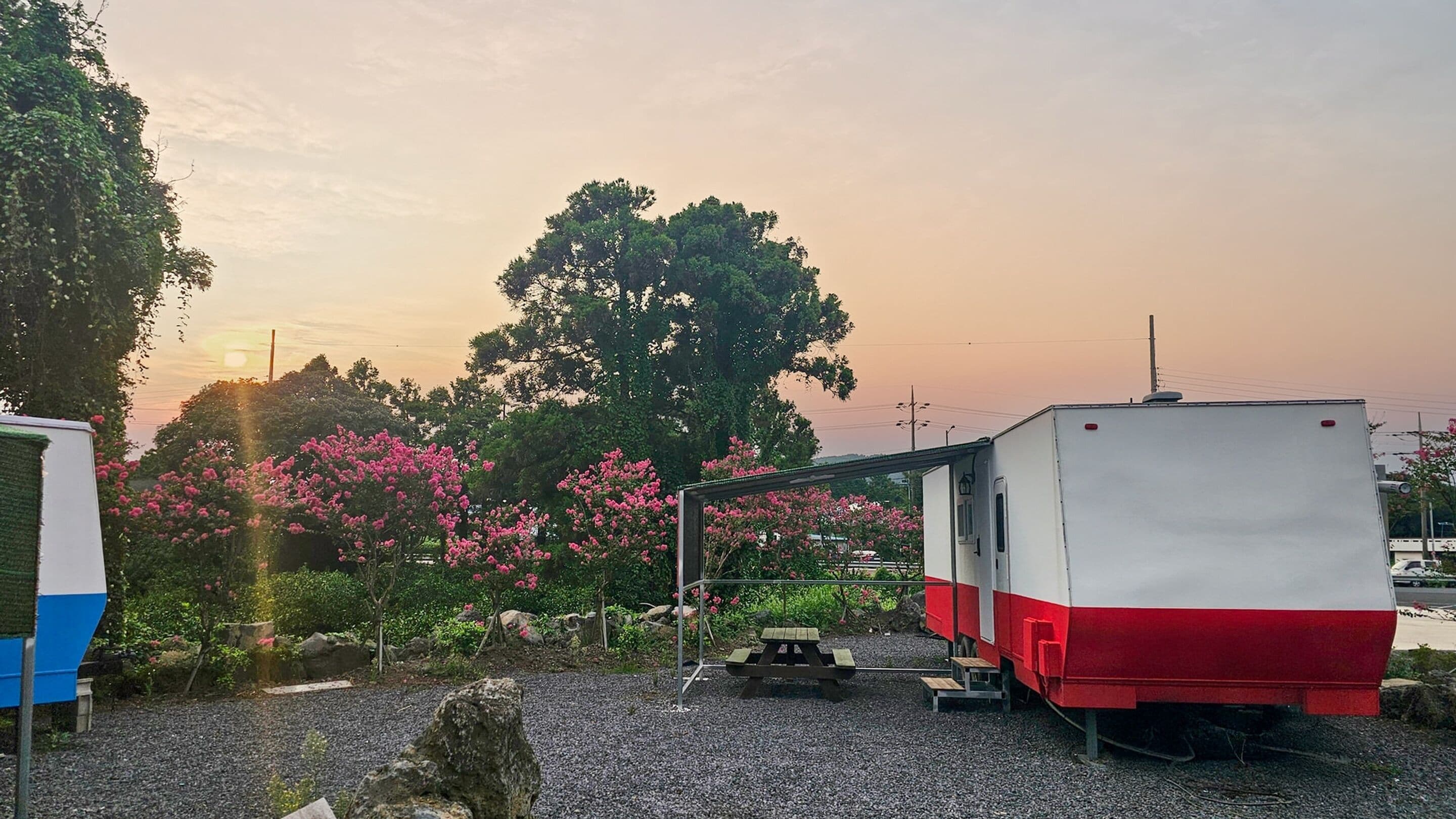 Una caravana estacionada, con diseño rojo y blanco, sobre superficie de grava, y una mesa de picnic cerca.