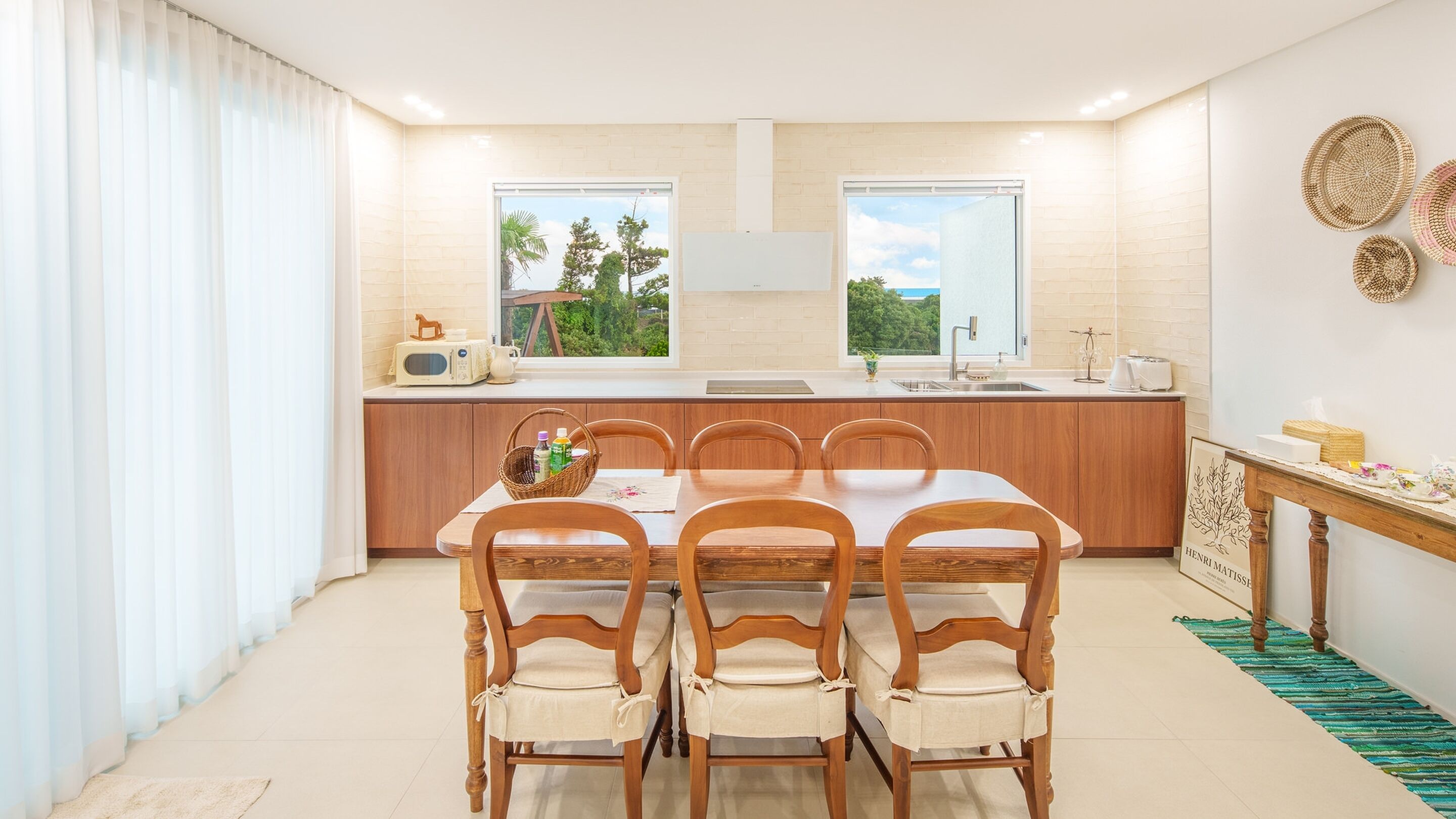 A modern kitchen with a dining area, featuring a wooden table and chairs, a built-in oven, and a view of greenery outside.