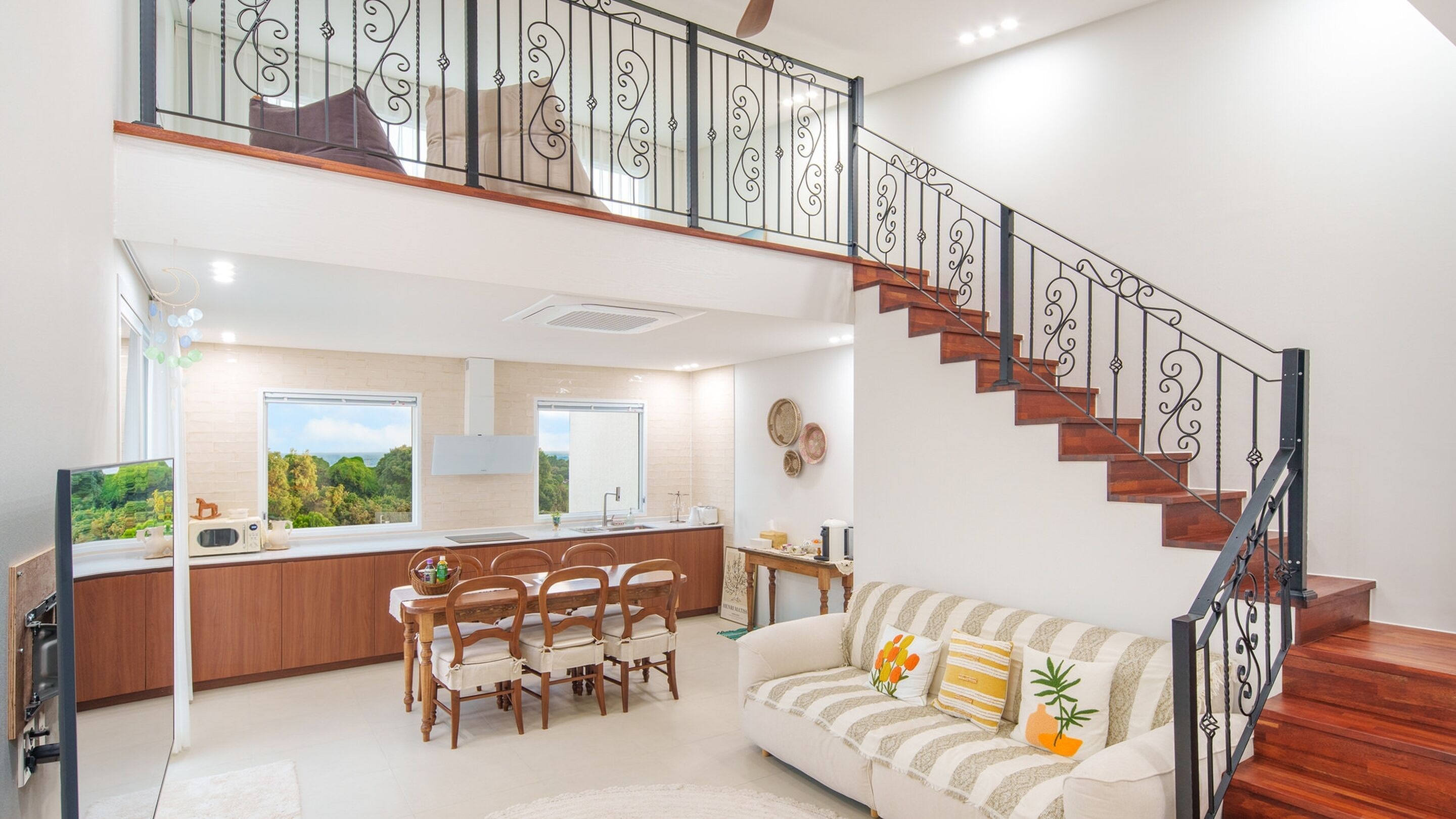 A spacious living area with a dining set, a sofa with decorative pillows, and a staircase with ornate railings.