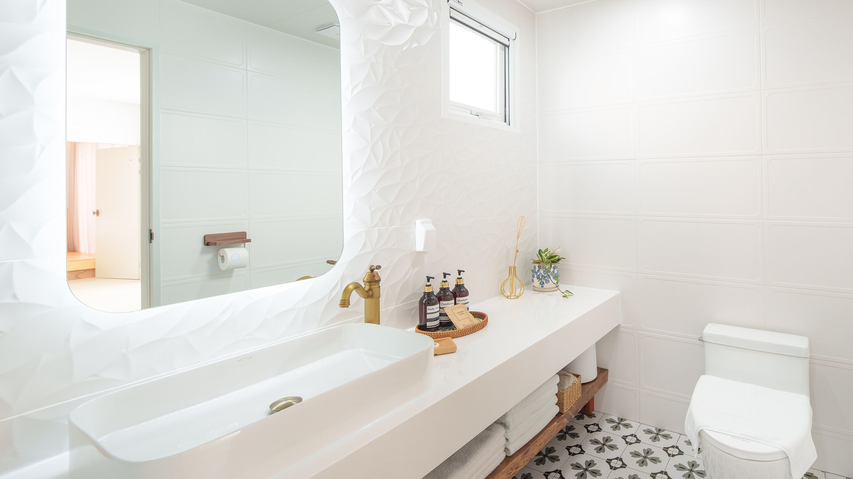 A modern bathroom with a large white sink, a gold faucet, and a white toilet. There is a mirror above the sink and a window above the toilet.