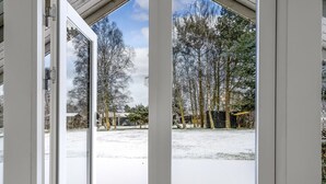 Vue depuis une pièce donnant sur une terrasse en bois qui mène à un espace extérieur enneigé, avec des arbres et un abri noir.