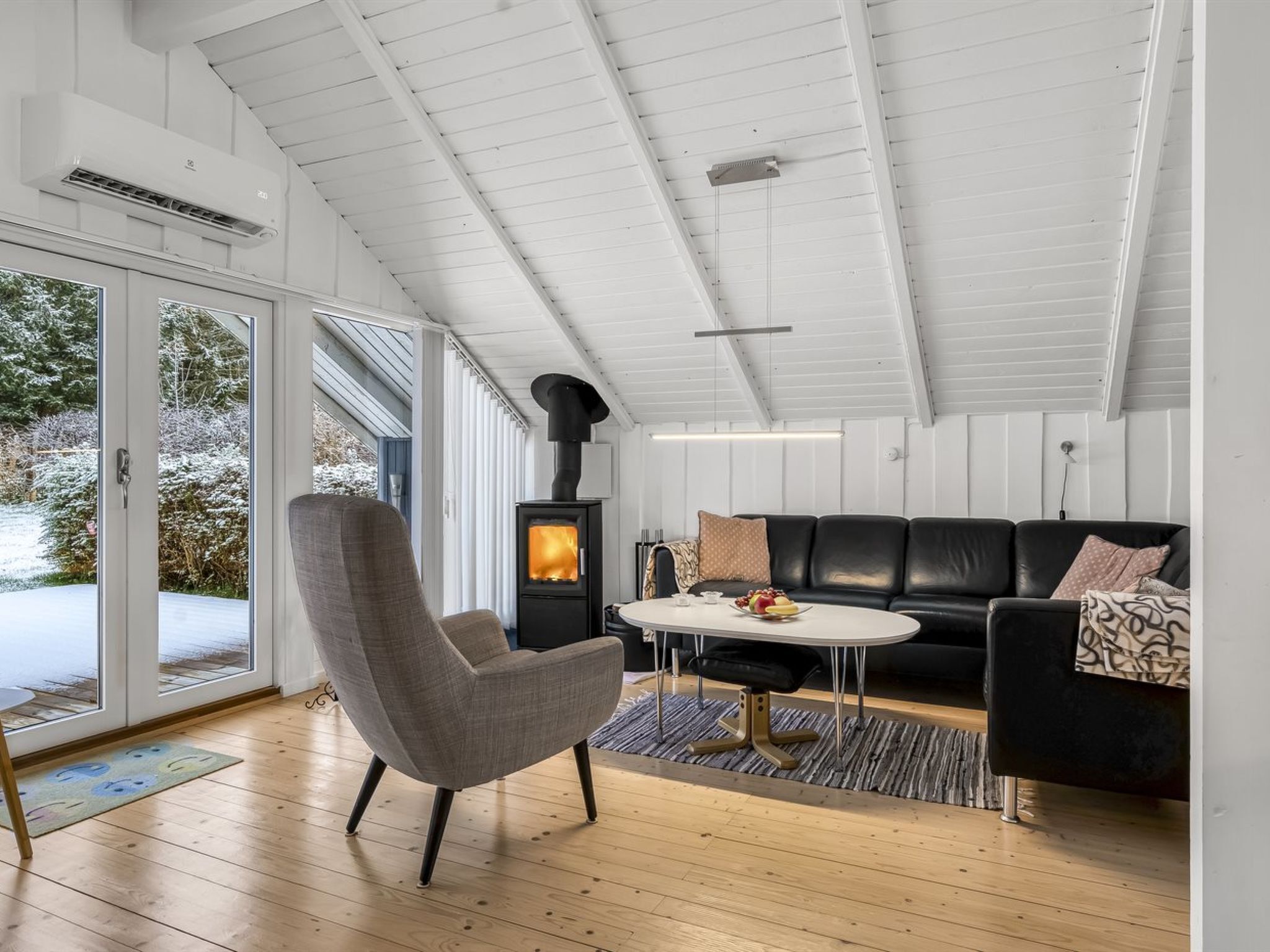 Un salon moderne avec un canapé en cuir noir, un fauteuil gris et une table basse ronde. On aperçoit un poêle à bois au fond de la pièce.