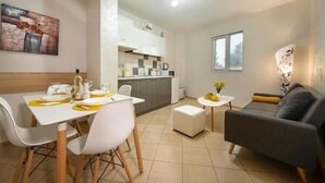 Un salon moderne avec un coin repas, une cuisine à l’arrière-plan et une grande fenêtre qui laisse entrer la lumière naturelle.
