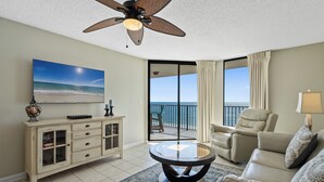 A living room with a ceiling fan, a large window with a view of the ocean, a beige sofa, a coffee table, and a cabinet with glass doors.