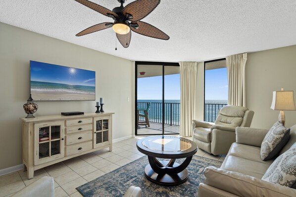 A living room with a ceiling fan, a large window with a view of the ocean, a beige sofa, a coffee table, and a cabinet with glass doors.