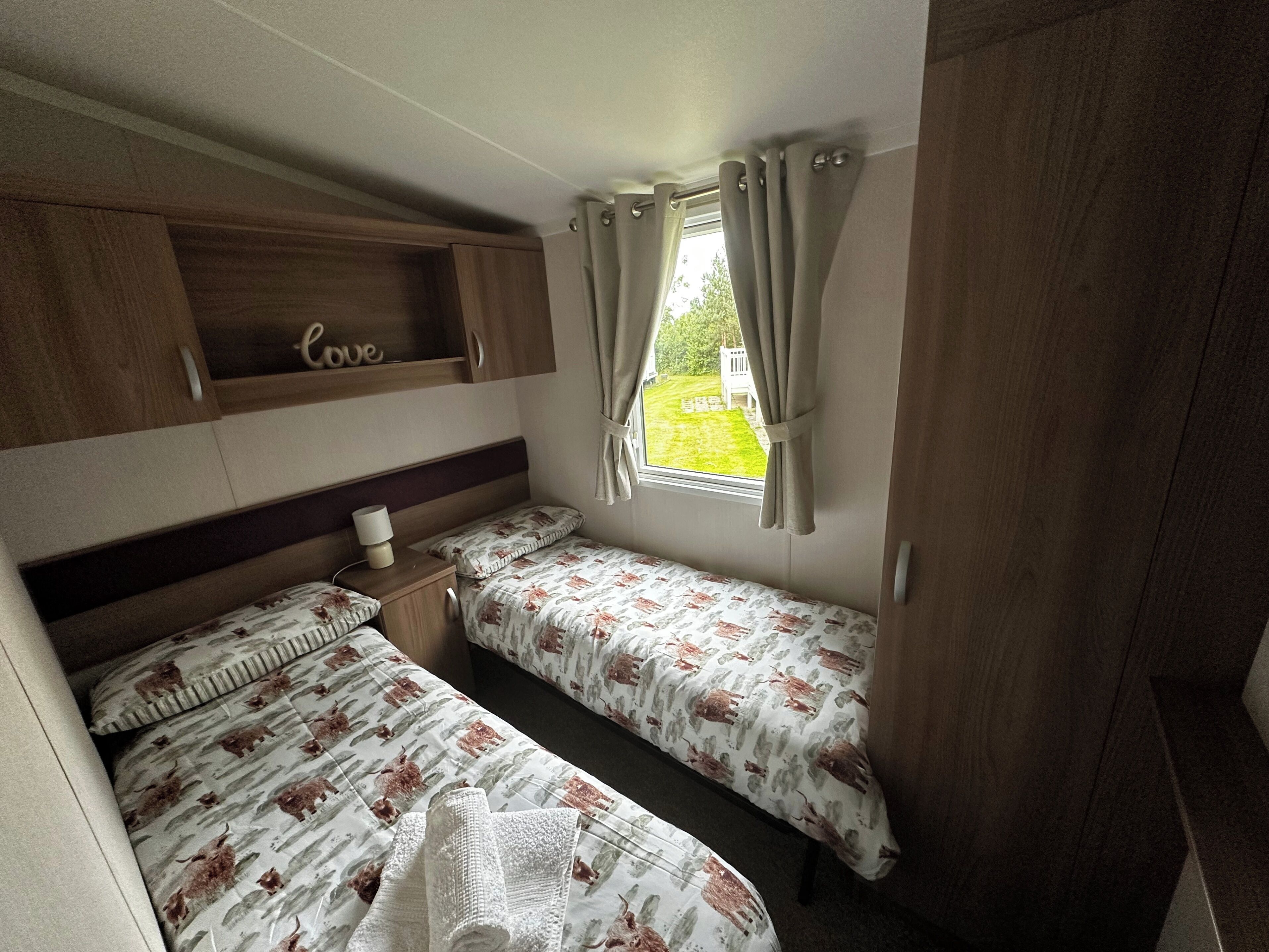 Ein kleines Schlafzimmer mit zwei Einzelbetten, einem kleinen Nachttisch, einer Ablage mit der Aufschrift 'love' und einem Fenster mit Vorhängen.