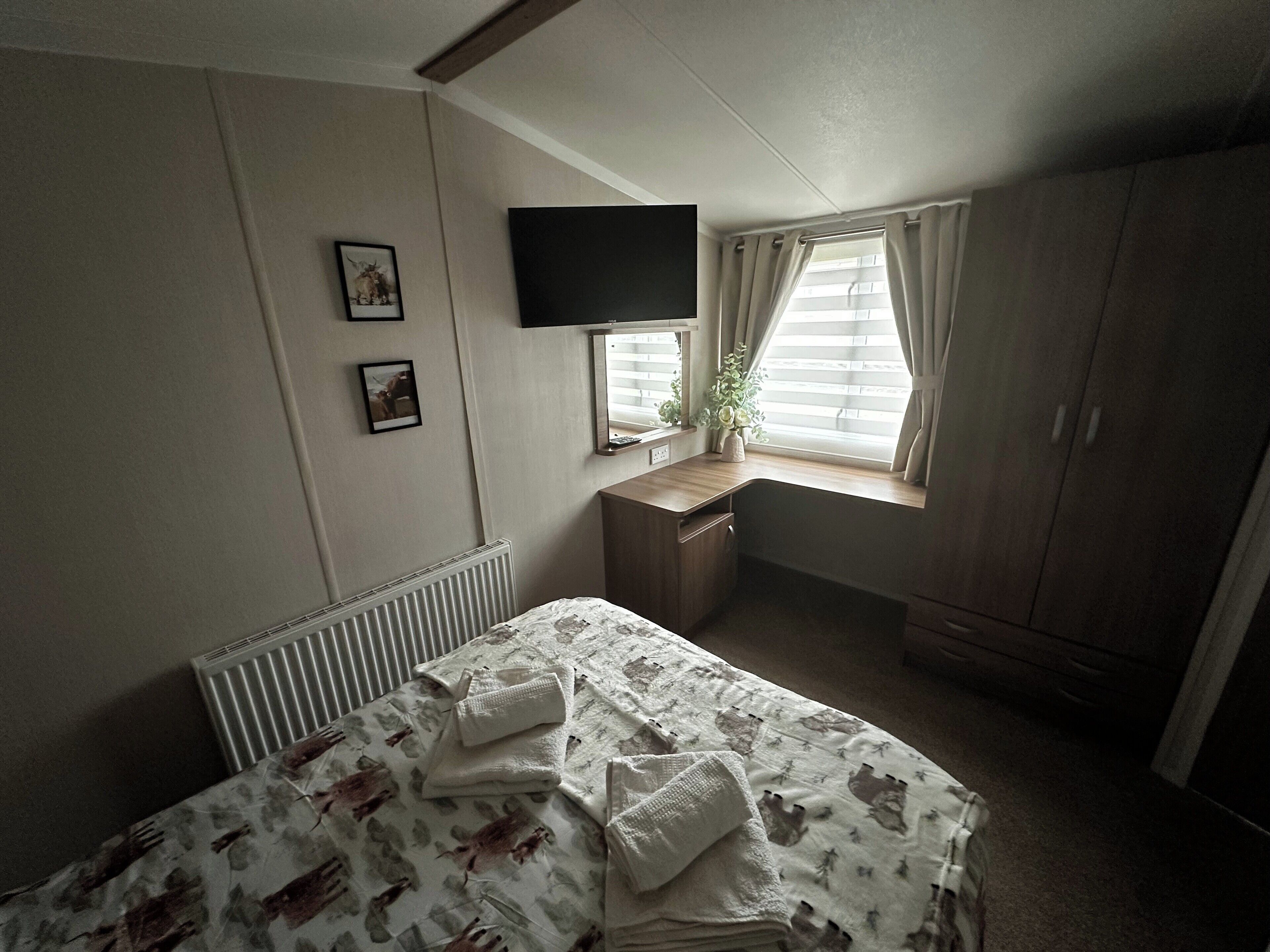 Quarto com cama, escrivaninha, televisão e janela com cortinas.