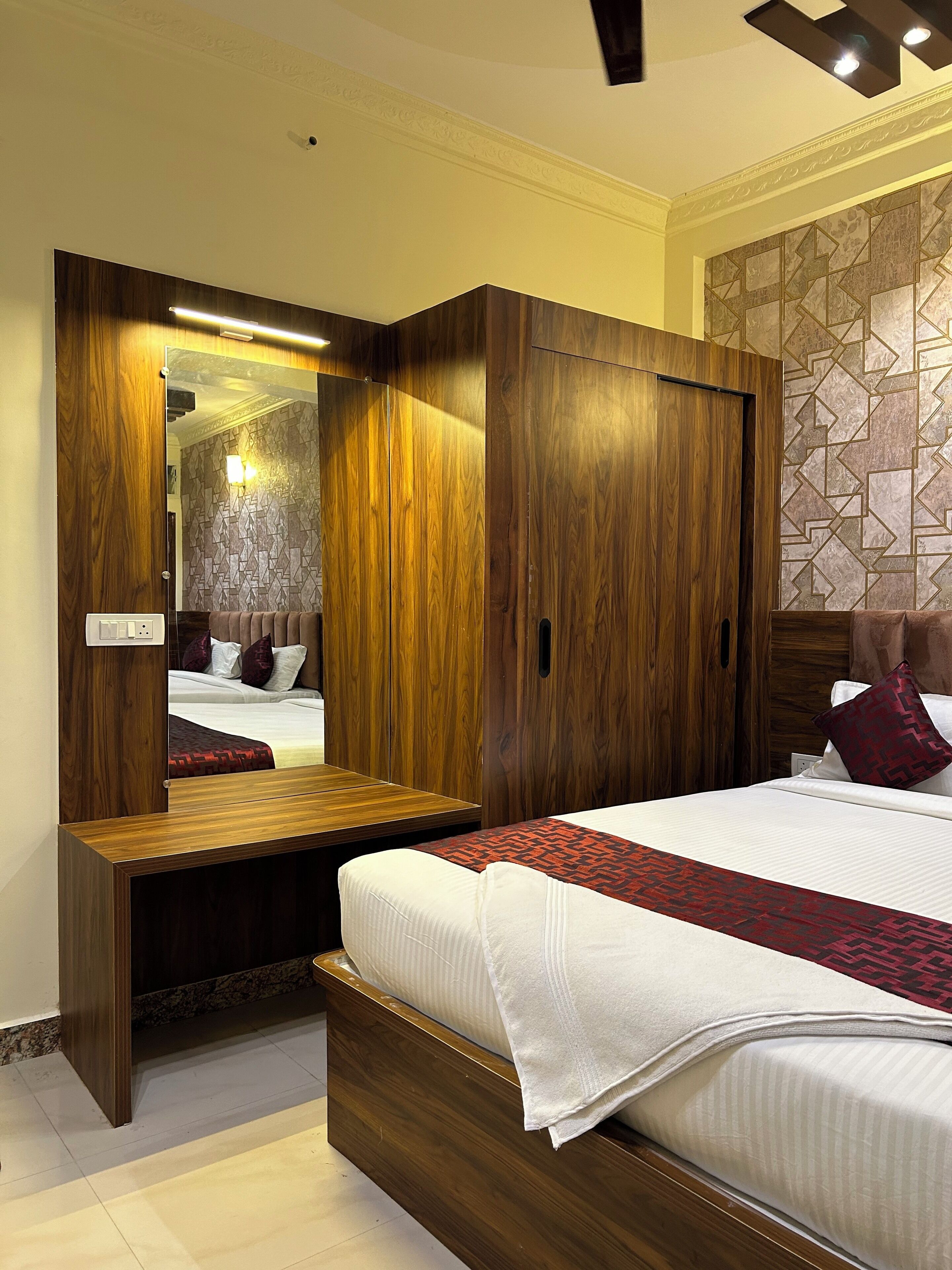 A bedroom with a wooden wardrobe, a bedside table, a bed with a patterned bedspread, and a wall with a geometric design.