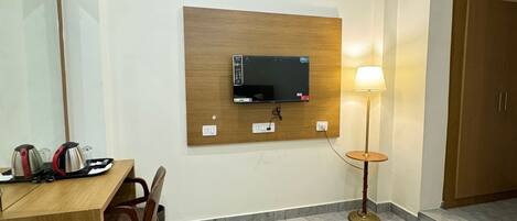 Una habitación de hotel con una cama, un escritorio con silla, televisor montado en la pared, una lámpara y un armario.