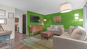 A living room with a green accent wall, a beige sofa, a wooden cabinet, and a desk with a lamp. - Quiet 2BR/2BA Olympia townhome on acreage near downtown, Capitol, workspace, garage parking (Olympia)