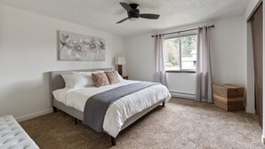 Ein Schlafzimmer mit einem Bett, einem Deckenventilator, einem Fenster mit Vorhängen, einem Wandbild und einem kleinen Korbwäscher.
