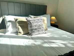 Una cama bien hecha con sábanas blancas, un cabecero tapizado en gris y una almohada estampada.