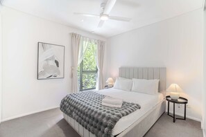 Une chambre à coucher comprenant un lit, deux tables de chevet, un ventilateur de plafond, une fenêtre avec des rideaux et un tableau abstrait accroché au mur.