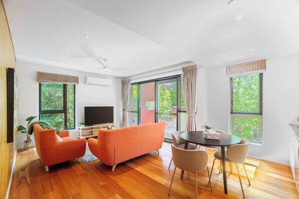 Una sala de estar moderna con televisor de pantalla plana, sofás anaranjados, una mesa de comedor con sillas y ventanas grandes con cortinas.