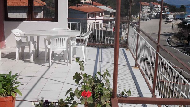 Un balcón con una mesa y sillas blancas, plantas en macetas y vistas de un pueblo costero.