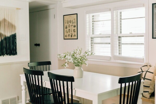Een eethoek met een witte tafel en zwarte stoelen, een vaas met bloemen en een raam met jaloezieën.