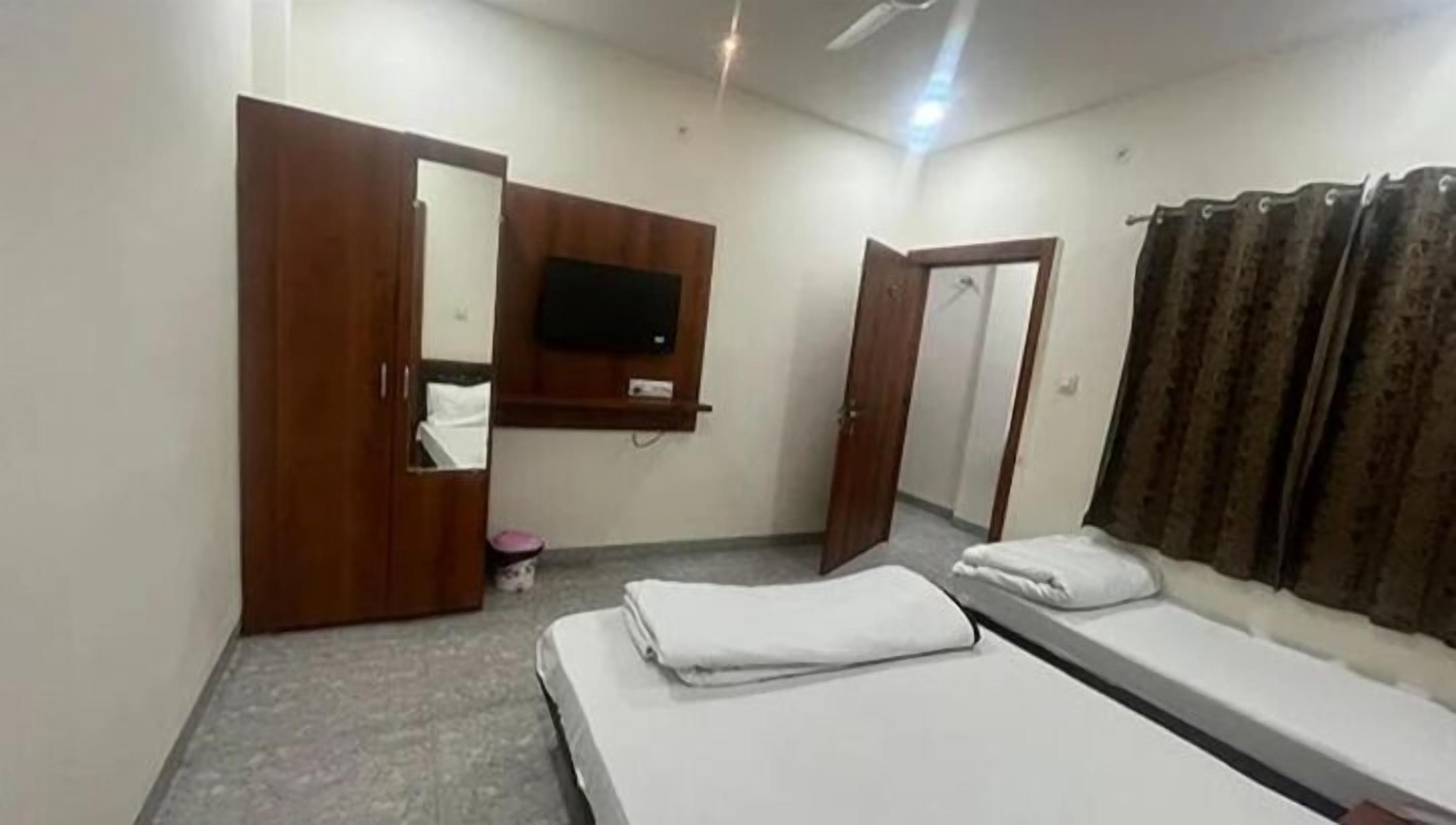A bedroom with two single beds, a wooden wardrobe, a television mounted on the wall, and a curtain on the window.