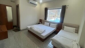 A bedroom with a large bed, a smaller bed, an air conditioning unit, a window with curtains, and a door. - Hotel Garima (Ujjain)