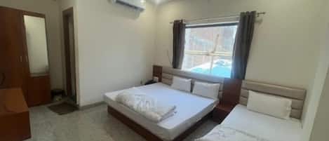 Une chambre à coucher avec un grand lit, un plus petit lit, un appareil de climatisation, une fenêtre avec des rideaux et une porte.