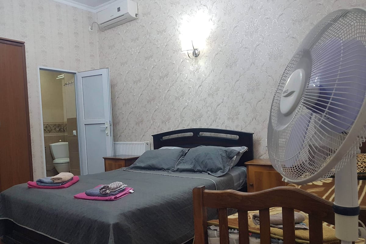 Une chambre à coucher avec un lit, une table de chevet, un ventilateur et une porte ouverte donnant sur une salle de bain.
