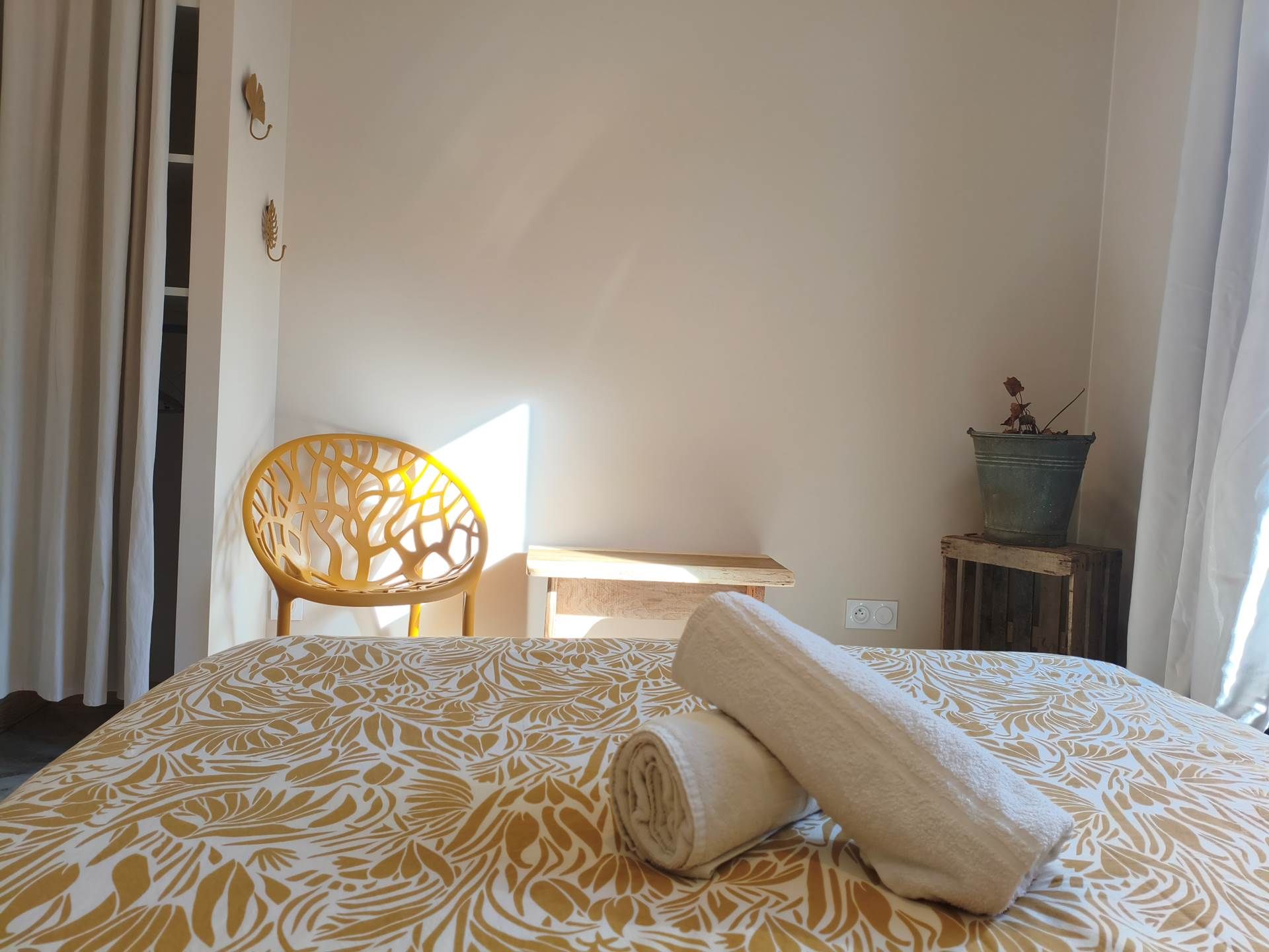 Ein ordentlich bezogenes Bett mit einem gemusterten Überwurf, ein zusammengerolltes Handtuch, ein Stuhl mit geometrischem Design, ein kleiner Tisch und ein Blumentopf.