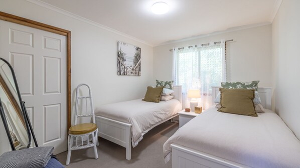 A bedroom with two single beds, a chair, a window with sheer curtains, and a door with a wooden frame. - Wanda Up, 80 Randall Dr - spectacular views, aircon and Wi-Fi (Salamander Bay)
